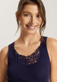 Top sin mangas azul marino con detalle de encaje floral en el escote, textura suave y tirantes anchos; mostrado en una persona sonriendo.