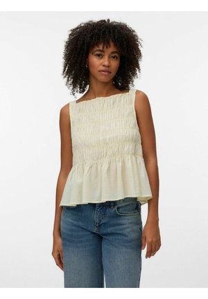 Femme portant un haut sans manches crème texturé avec un ourlet volanté et un jean bleu sur un fond blanc uni.