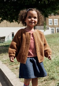 Brun, blød fleecejakke, rød- og hvidstribet skjorte, denimnederdel og farverige sokker. Barn står udenfor med græs og træer.