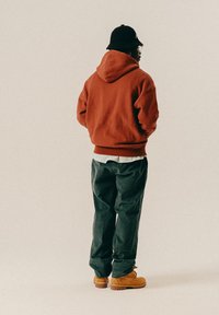 Person som står med ryggen til iført en rustfarget hettegenser, mørkegrønne bukser, svart bøttehatt, briller og lyse arbeidssko, på en ensfarget bakgrunn.