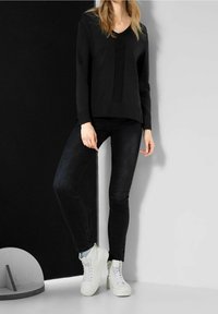 Czarny dzianinowy sweter z dekoltem w serek i teksturowaną wstawką, w połączeniu z czarnymi obcisłymi dżinsami i białymi sneakersami za kostkę. Tło w ciemnych kolorach.