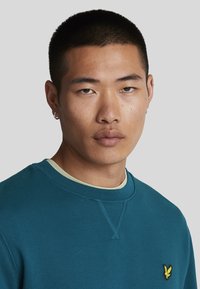 Sudadera de cuello redondo en color azul verdoso con un logo amarillo bordado en el lado izquierdo del pecho, con cuello acanalado y un acabado suave y texturizado.