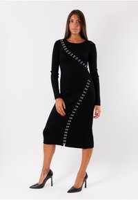 Abito nero a coste con maniche lunghe, dettagli asimmetrici nei bottoni con accenti in argento, taglio al ginocchio. Abbinato a tacchi neri.