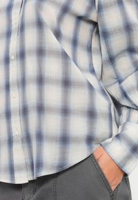 Camisa leve de xadrez nas tonalidades branca, azul e cinza. Apresenta fecho por botões e mangas longas com um hem arredondado. Tecido suave e transparente.