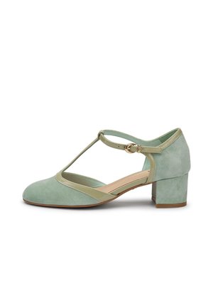 Lichtgroene suède dames T-strap schoen met lage blokhak, beige afwerking en gouden gesp op een witte achtergrond.