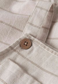 Gros plan de tissu beige avec un motif à carreaux marron clair, comportant un bouton en bois et un détail de sangle cousu.