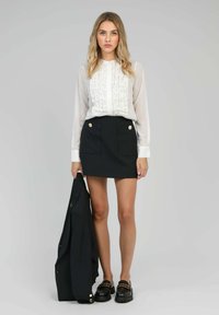 Femme portant un chemisier blanc à volants et une mini-jupe noire, tenant un blazer noir avec des mocassins noirs épais, sur un fond gris clair.