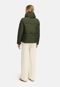 Woman standing with back facing camera, wearing an olive green puffer jacket, cream wide-leg pants, and beige sneakers.