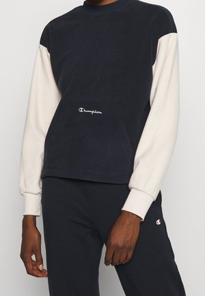 Persoon draagt een marineblauw-wit Champion sweater met lange mouwen en bijpassende marineblauwe sweatpants, staand tegen een effen achtergrond.