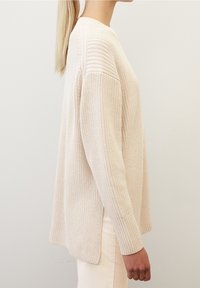 Maglione lavorato a maglia in un morbido beige, con una texture a costine, maniche lunghe e un orlo posteriore leggermente più lungo con spacchi laterali per una migliore ventilazione.