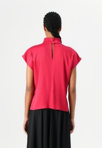 Red short-sleeve blouse with a high collar, featuring a keyhole opening at the back, made from a smooth, lightweight fabric.