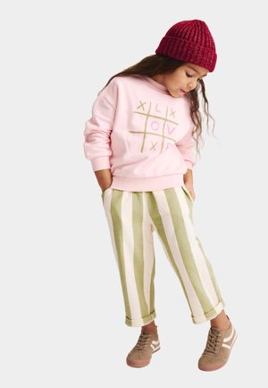 Lichtroze sweatshirt met goud- en roze grafisch ontwerp, gecombineerd met crèmekleurige en groene gestreepte broek en bruine sneakers. Rode gebreide muts op het hoofd.