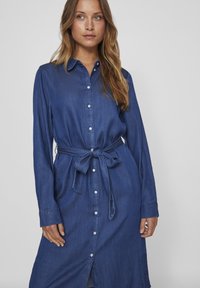 Denim srajčna obleka v temno modri barvi z dolgimi rokavi, zavezovanjem v pasu in odprtino spredaj. Vključuje bele gumbe in ovratnik.