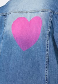 Chaqueta de mezclilla con un gran diseño de corazón rosa en la parte posterior. La tela es de color azul claro, con una textura suave y detalles de costura visibles.