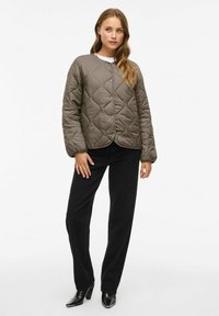 Femme portant une veste matelassée olive, un pantalon large noir et des bottes à talons noires, se tenant devant un fond blanc uni.