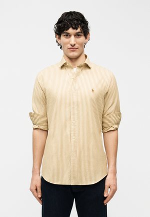 Homme aux cheveux bouclés portant une chemise à rayures verticales beige et blanche à boutons, manches retroussées, et un pantalon sombre, debout devant un fond blanc.