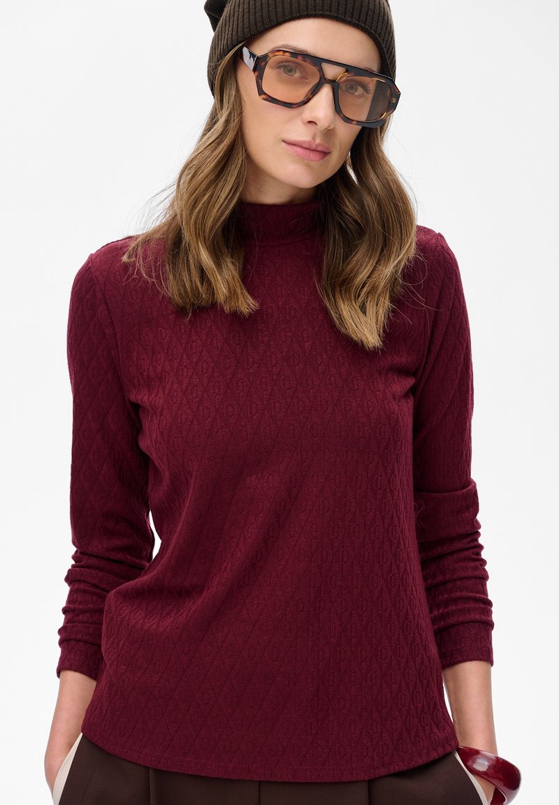 Kobieta ubrana w bordową, textured koszulę z długim rękawem, brązową czapkę, duże okulary w skorupie żółwia oraz bordo bransoletkę, z rękami w kieszeniach.