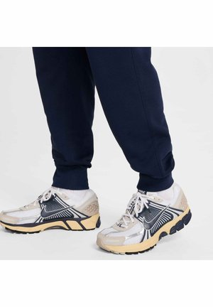 Baskets de sport blanches et beiges avec des accents noirs et des panneaux en maille, associées à un pantalon de jogging bleu marine au niveau de la cheville.