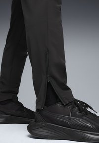 Sorte træningsbukser med lynlås ved anklerne, parret med sorte sneakers. Stoffet er glat, med en tætsiddende design der fremhæver benet.