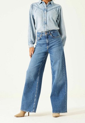 Kobieta ubrana w jasnoniebieską dżinsową koszulę zapinaną na guziki z bufkami na ramionach oraz wysokowtalowane szerokie niebieskie dżinsy, widoczne beżowe buty na obcasie.