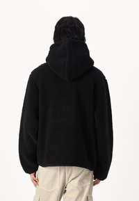 Felpa nera in fleece con cappuccio strutturato, vestibilità over, e superficie testurizzata, abbinata a pantaloni cargo beige. Solo vista da dietro.