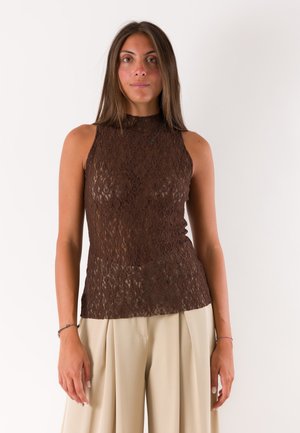 Donna con capelli castani lisci indossa un top smanicato in pizzo marrone e pantaloni larghi beige chiaro su sfondo bianco semplice.