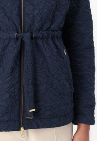 Σακάκι ναυτικό μπλε, με υφή, ελαστική μέση με κορδόνι, μπροστινό φερμουάρ και χρυσά αξεσουάρ. Διαθέτει σχέδιο από τσαλακωμένο ύφασμα.