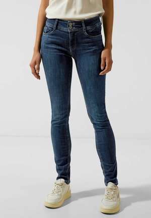 Vrouw die donkerblauwe slim-fit jeans en witte sneakers draagt, staand tegen een effen achtergrond.