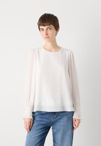 Witte blouse met lange doorschijnende mouwen en ruches aan de manchetten; heeft een gladde, lichte stof en een ronde halslijn. Gecombineerd met blauwe jeans.