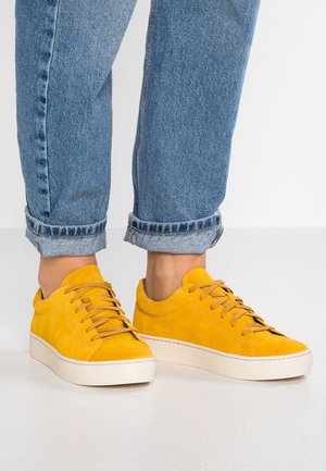 Gule ruskindssneakers med lysebrune snørebånd, båret med opkørte blå cowboybukser på en hvid overflade.