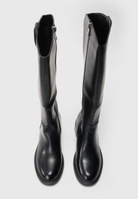Une paire de bottes noires en cuir jusqu'au genou avec des zips sur le côté et des orteils ronds, posées verticalement sur un fond clair.