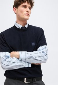 Νέος άνδρας με σγουρά μαλλιά που φοράει μπλε πουλόβερ πάνω από μπλε πουκάμισο με ρίγες, με σταυρωμένα τα χέρια, κοιτάζοντας ελαφρώς προς τα δεξιά.