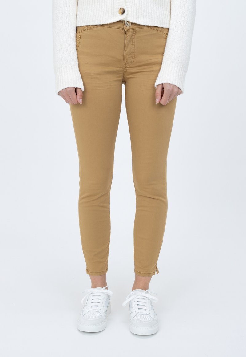 Jeans skinny color caqui con una textura suave, con cierre de cremallera y botón, combinados con zapatillas blancas y un suéter de canalé color crema.