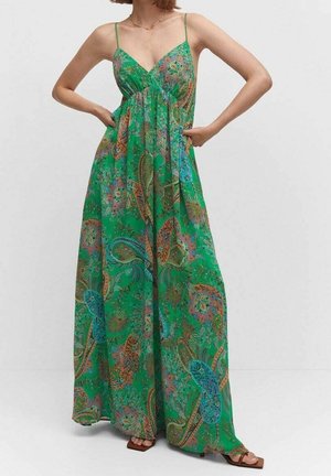 Kobieta w zielonej, bezrękawnikowej sukience maxi z kolorowymi wzorami paisley, stojąca z rękami na biodrach, ubrana w brązowe sandały na obcasie z odkrytymi palcami.