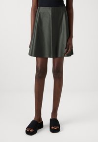 Una gonna in ecopelle verde scuro con un design svasato, che si estende sopra il ginocchio, abbinata a sandali platform neri.