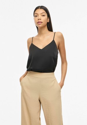 Kvinde med langt mørkt hår iført en sort ærmeløs V-hals top og højtaljede beige bukser med hænderne i lommerne, foran en hvid baggrund.