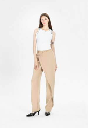 Joven mujer con cabello largo viste una blusa blanca sin mangas y pantalones anchos beige con panel frontal superpuesto, de pie frente a un fondo blanco.