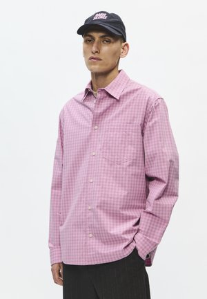 Homme portant une chemise ample à carreaux roses boutonnée, un pantalon noir et une casquette noire avec le texte "Radio Silence" sur fond blanc uni.
