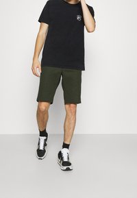 Černé bavlněné tričko s logem na přední straně, zelené šortky s texturovaným povrchem a černé sportovní boty s bílými tkaničkami.