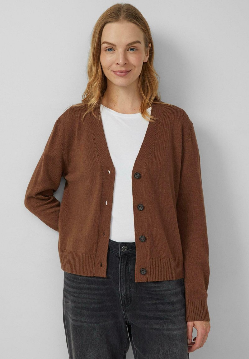 Brauner Cardigan in Strickstruktur mit V-Ausschnitt und sechs schwarzen Knöpfen. Eng anliegende Bündchen und Saum, getragen über einem weißen Hemd und kombiniert mit dunklen Jeans.