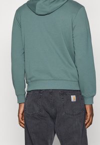 Mikina s kapucí v tlumené tyrkysové bavlně s žebrovanými manžetami a lemem, doplněná tmavě šedými džínami s etiketu na zadní kapse.