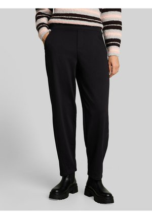 Person trägt schwarze gerade Hose, schwarze klobige Stiefel und einen gestreiften Pullover mit teilweise in den Taschen steckenden Händen.