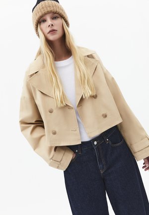 Ung kvinde med langt blondt hår iført en beige cropped trenchcoat, hvid skjorte, mørke jeans og en strikket beige hat.