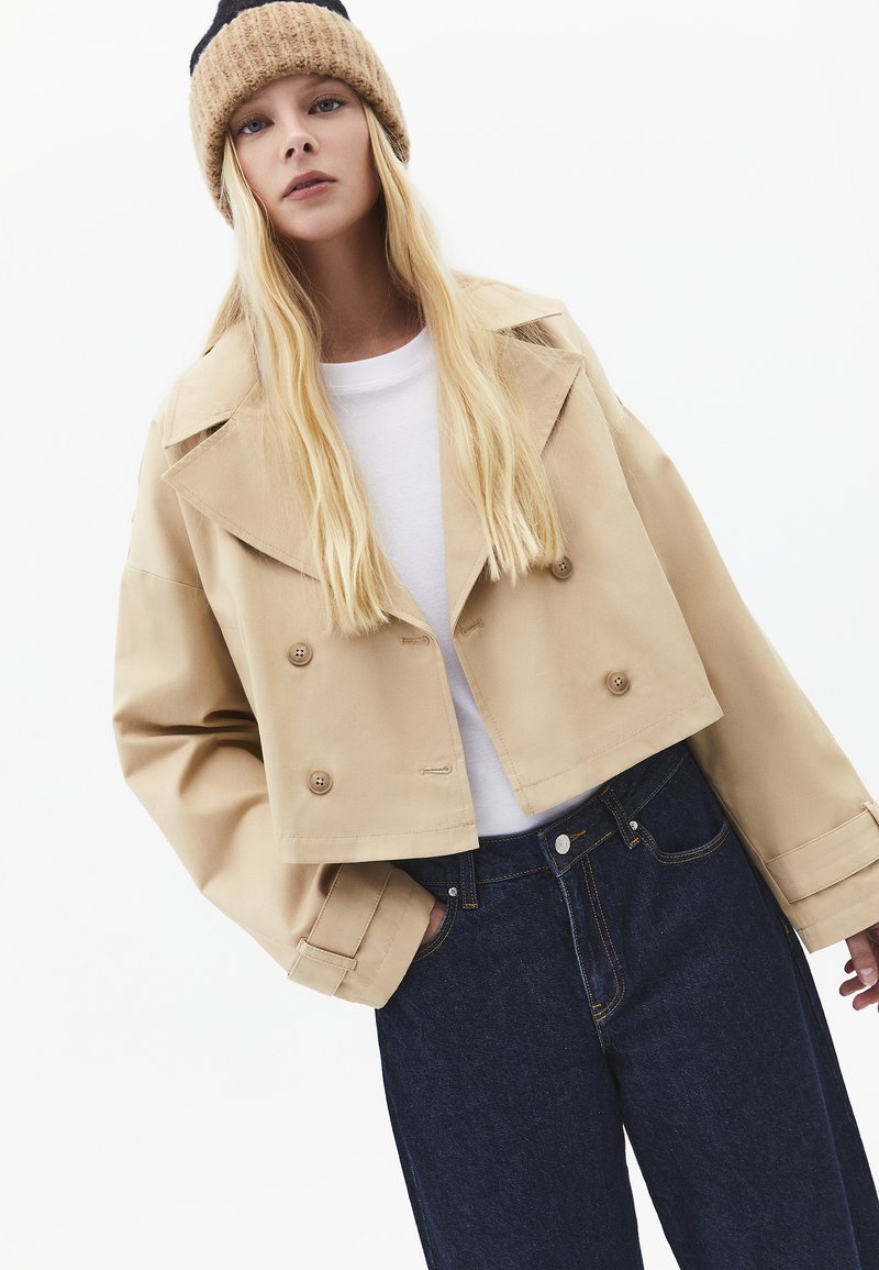 Mladá žena s dlouhými blond vlasy, oblečená v béžovém krátkém trenčkotě, bílé košili, tmavých džínách a pletené béžové čepici.