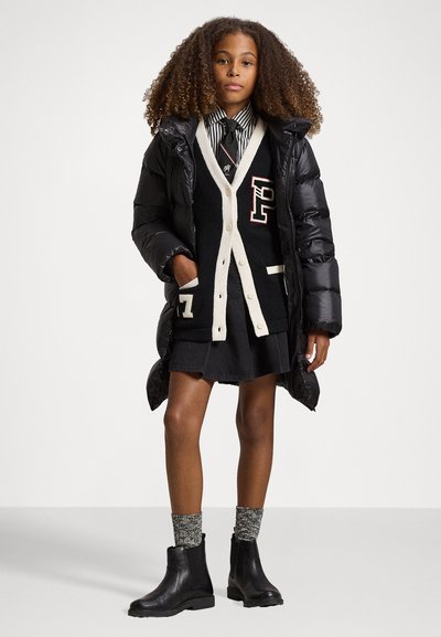 Fekete пухи kabát fekete és krémszínű logós kardigán felett, farmer szoknya és textrált kötött zoknis bokacsizmák.