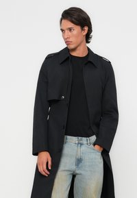 Zwarte trenchcoat met een grote kraag, knoopsluiting aan de voorkant en schouderaccenten, gedragen over een zwart shirt en lichtblauwe jeans.