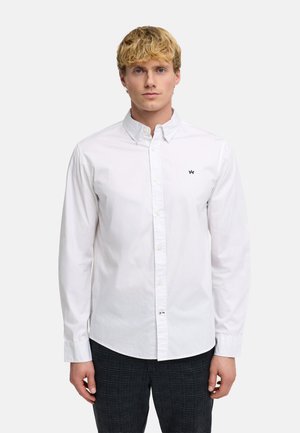 Lyshåret mand iført hvid skjorte med knapper og lille sort logo på brystet, kombineret med mørke ternede bukser, stående mod en ensfarvet baggrund.