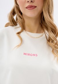 Crème sweatshirt met roze geborduurde tekst "MIRONS" op de voorkant, gecombineerd met een parelketting. Soepele stof, relaxte pasvorm, ronde halslijn.