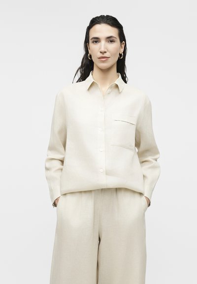 Γυναίκα με σκούρα, βρεγμένα μαλλιά φοράει χαλαρό λευκοκρεμ πουκάμισο με κουμπιά και μακρυά μανίκια, και παντελόνι φαρδύ στο χρώμα, με τα χέρια στις τσέπες, ουδέτερο φόντο.