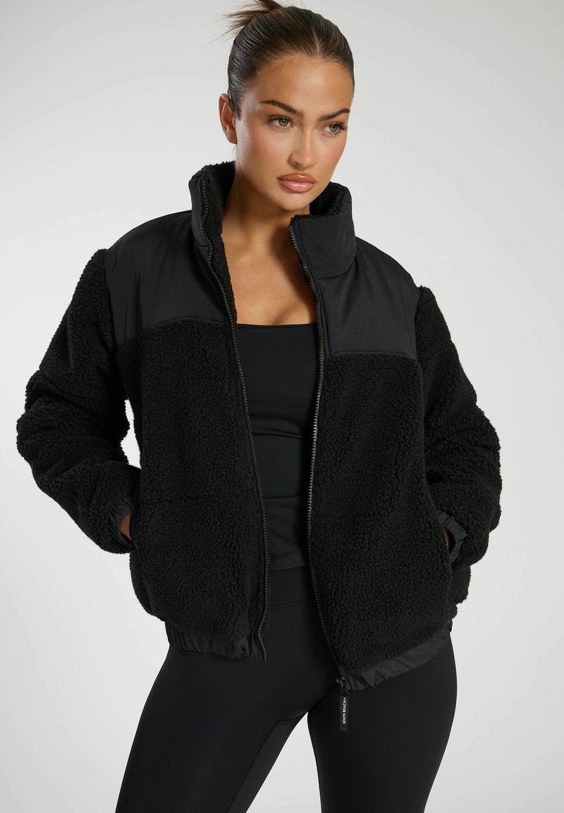 Schwarze Zip-Jacke mit einem strukturierten Fleece-Oberteil, glatten schwarzen Ärmeln, hohem Kragen und elastischem Saum. Schlichtes Design mit seitlichen Taschen.